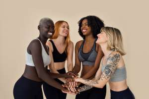 Happy diverse fit sporty young women, multicultural models in sports wear stand in circle at beige background join hands together in pile stack advertising yoga group gym trainings, team workout.