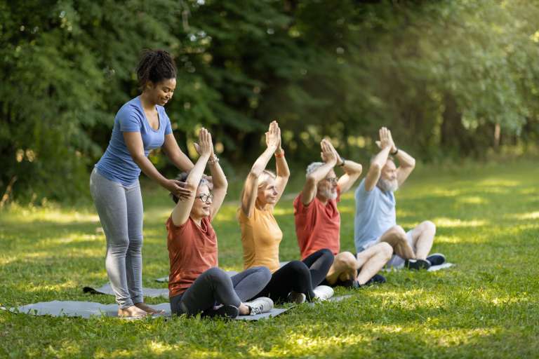 Outdoor Yoga Class. Group of smiling senior people training together outdoors, happy mature men and women in sportrswear sitting in lotus position with hands above head, enjoying healthy lifestyle